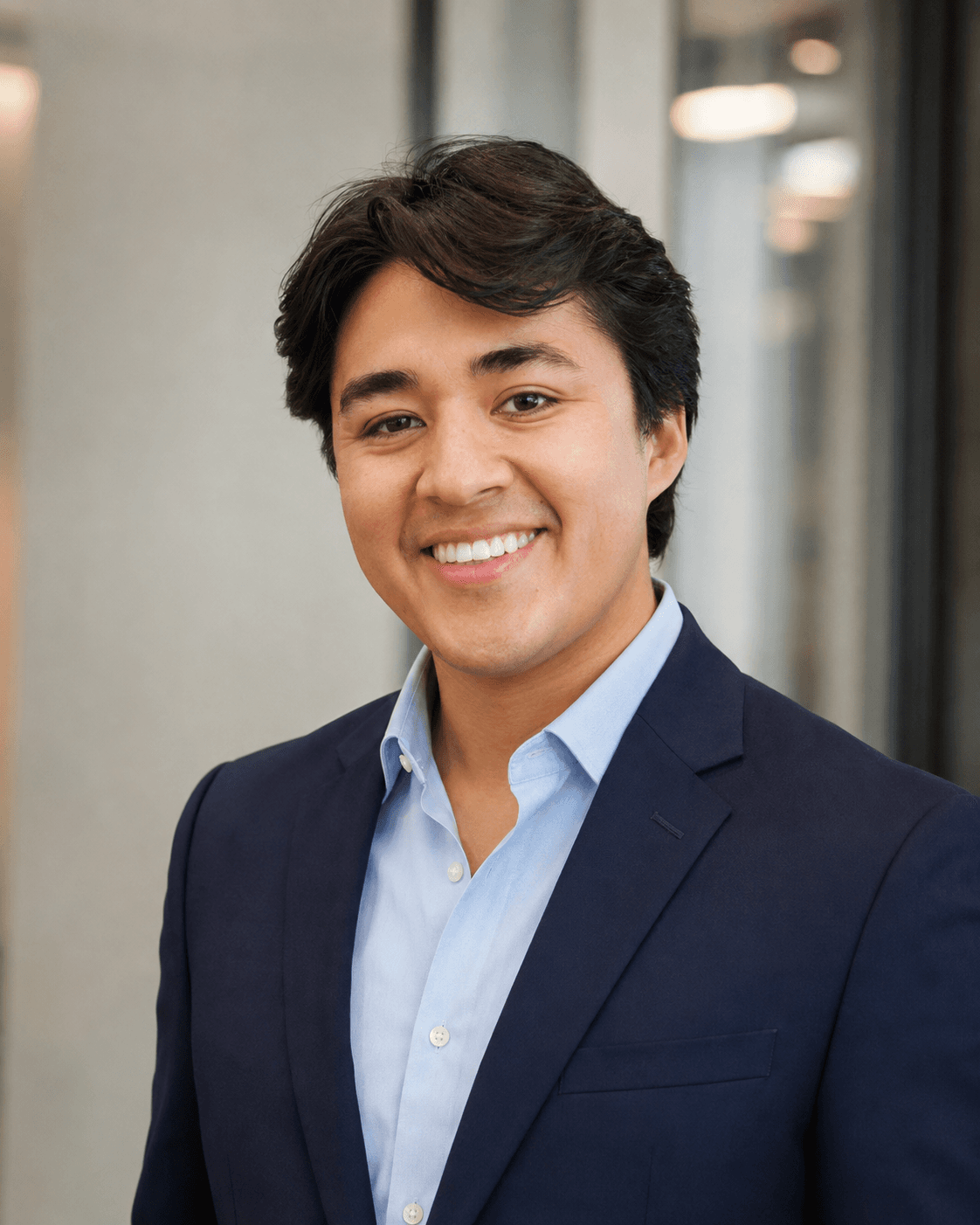 Smiling young man with dark wavy hair wearing a navy blazer and light blue shirt.